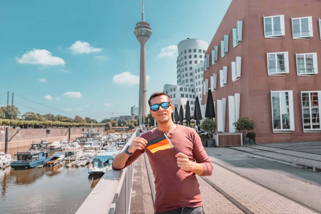 A young happy man with a German flag poses at the Media Harbor and TV-tower in Dusseldorf, illustrating the concept of studying language abroad and traveling while benefiting from FEIE.
